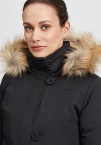 Manteau d'hiver noir avec une capuche en fourrure, doté de grands boutons, d'une surface texturée et de poches latérales.