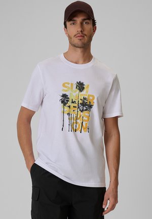 Mężczyzna w białej koszulce z żółtym napisem "SUMMER SEASON" i czarnymi grafikami palm, czarne spodnie i brązowa czapka na szarym tle.