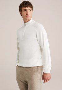 Weißer Strickpullover mit einem Viertelreißverschluss-Kragen, zeichnet sich durch Raglanärmel, gerippte Bündchen und eine glatte Textur aus. Kombiniert mit hellbraunen Hosen.