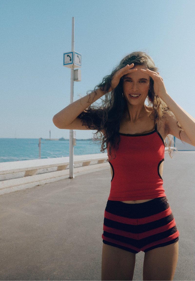 Femme en débardeur rouge et short rayé, se protégeant les yeux avec les mains près d'une promenade au bord de la mer sous un ciel dégagé.