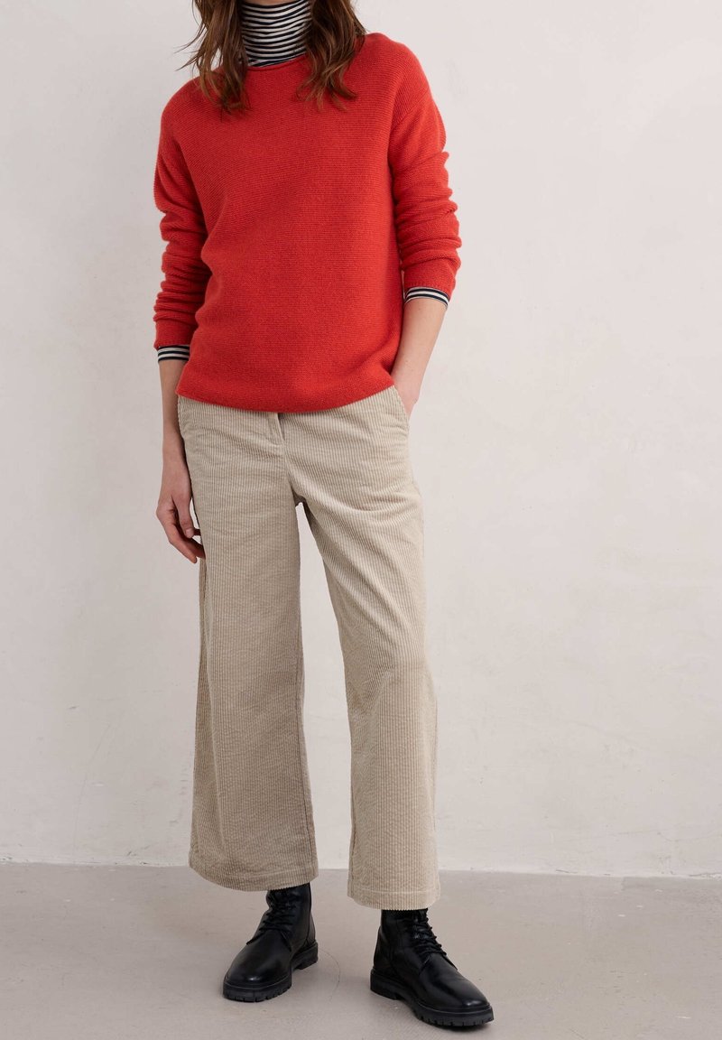 Femme en pull rouge, col roulé rayé, pantalon beige large en velours côtelé et bottes noires à lacets debout contre un mur clair uni.
