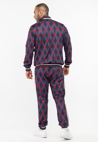 Marineblauwe en rode tracksuit met ruitpatroon, voorzien van een ritskraag, gestreepte manchetten en een elastische tailleband. Witte sneakers maken de look compleet.