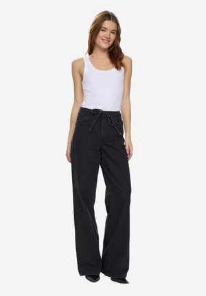 Tânără purtând un top alb fără mâneci și pantaloni largi negri cu talie înaltă, zâmbind cu o postură relaxată.