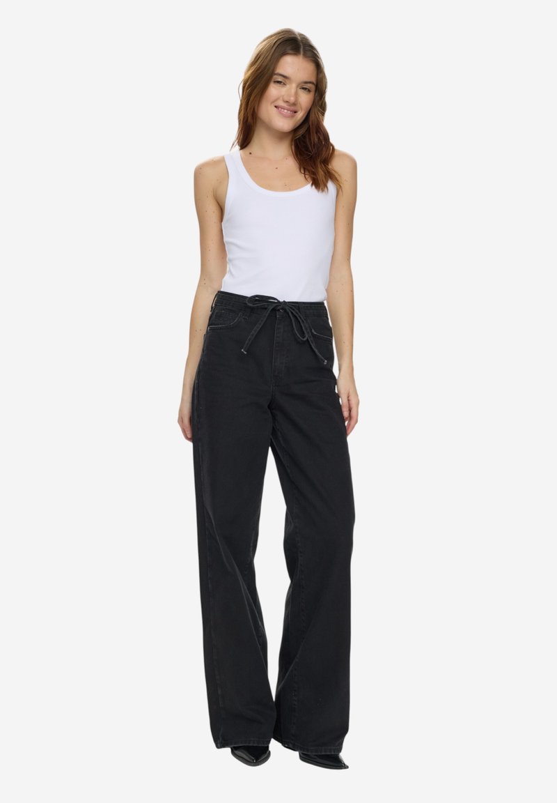 Jeune femme portant un top blanc sans manches et un pantalon large noir taille haute, souriante avec une posture détendue.