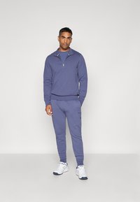 Muž stojaci v zhodujúcom sa fialovom sveteri s krátkym zipsom a teplákových nohaviciach, s bielymi teniskami na pozadí svetlej farby.