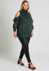 Grön tryckt blus med kalla axlar, volangärmar och hög krage, parat med svarta leggings i konstläder och svarta klacksandaler.