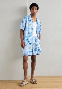 Ensemble composé d'une chemise à manches courtes bleu clair et d'un short, décoré de motifs de palmiers et de voitures. Porté avec un débardeur blanc et des sandales marron.
