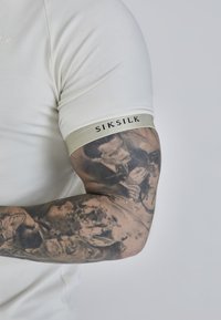 Valkoinen lyhythihainen paita, jossa on harmaa hihansuukuva ja johon on kirjailtu "SIKSILK" mustalla. Hihassa näkyy monimutkaisia tatuointeja, joissa on yksityiskohtaisia kuvioita.
