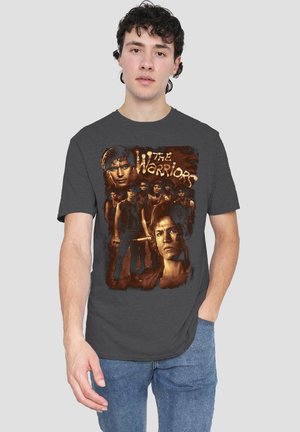 Junger Mann trägt ein dunkelgraues T-Shirt mit einer Grafik von mehreren Männern und dem Text "Die Krieger" in stilisierten Buchstaben.
