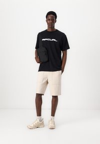 Fekete póló fehér logóval, krémszínű rövidnadrág és bézs sportcipő. Könnyű, fekete crossbody táska. A modell egy fehér háttér előtt áll.