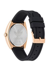 Montre avec bracelet en silicone noir et boîtier en acier inoxydable doré rose. Présente un fond argenté gravé du logo et un cadran de forme ronde.