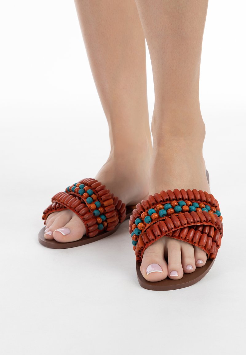 Sandali piatti marroni con accenti di perline rosse e blu intrecciate. Il design presenta una punta aperta ed è realizzato con materiali morbidi.