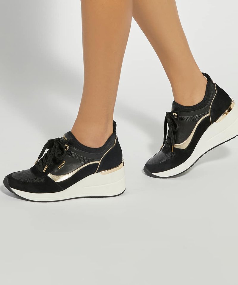 Jambes portant des baskets compensées noires et dorées avec des semelles blanches, debout sur une surface grise claire unie.