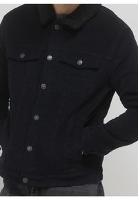 Homme portant une veste en velours côtelé noire avec un col en peau retournée noire et des boutons-pression en métal, les mains dans les poches latérales, pantalon gris visible en dessous.