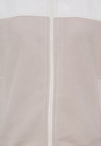 Veste polaire beige et blanche avec fermeture éclair à l'avant et deux poches latérales, montrant un tissu doux et texturé et des coutures nettes.