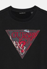 Felpa nera con un patch triangolare in tessuto grigio e un logo ricamato rosso con punto interrogativo, in contrasto con lo sfondo uniforme.