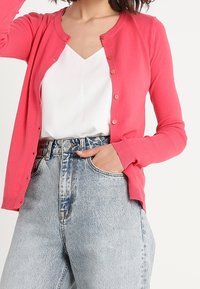 Pink knitted cardigan with round neckline and button closures, worn over a white top, paired with high-waisted light blue jeans.