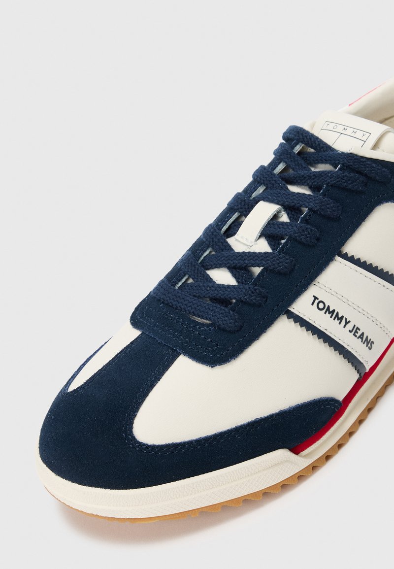 Baskets blanches et marines avec des lacets marins, logo "Tommy Jeans" sur le côté, et une semelle en gomme, légèrement inclinées pour montrer le bout et le côté.