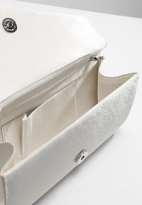 Bolso de mano de satén blanco con cierre magnético, abierto para mostrar el bolsillo interior y la sujeción de la cadena.
