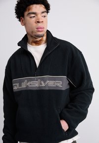 Sort fleece pullover med en kvart-zip, har en grå vandret stribe med "Quiksilver" logo, tekstureret overflade og sidelommer.