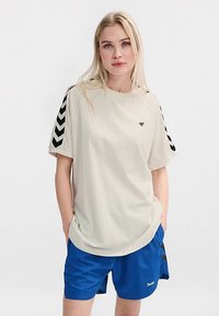 Lichtbeige T-shirt met korte mouwen en zwarte chevronstrepen op de mouwen. De stof is glad en heeft een klein logo op de borst.