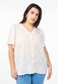Blusa crema chiaro con pattern floreale testurizzato, scollatura a V e maniche corte. Presenta bottoni lungo il davanti e una vestibilità ampia.
