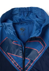 Blauer Windbreaker mit Kapuze, ausgestattet mit einem großen rot akzentuierten "S"-Logo, Reißverschluss und glatter, glänzender Textur.