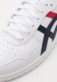 Vit lädersneaker med texturerad yta, har vita snörningar, marinblå och r öda accenter, samt perforerad detaljering på tårna.
