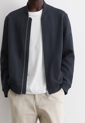 Homme portant une veste bomber bleu marine zippée sur un t-shirt blanc et un pantalon beige, debout devant un fond blanc uni.