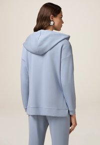 Felpa con cappuccio azzurro chiaro dal taglio rilassato, texture morbida e polsini a costine. Presenta una tasca a marsupio e un fondo posteriore più lungo.