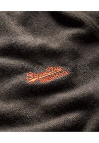 Dunkelbrauner Stoff mit einer weichen Textur, der ein auffälliges, orangefarbenes, gesticktes "SuperDry"-Logo zeigt. Die Oberfläche zeigt ein dezentes, gewebtes Muster.