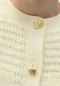 Kremowy dzianinowy kardigan z teksturowanymi panelami, ozdobiony dwoma dekoracyjnymi złotymi guzikami w kształcie splecionych kół.
