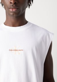 Balta be rankovių marškinėliai su apvaliu kaklu, su siuvinėjamu "Calvin Klein Jeans" logotipu smėlio ir šviesiai rudos spalvos krūtinėje. Minkšta medvilnės tekstūra.