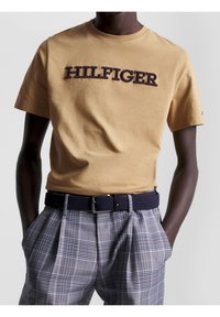 Man som har på sig en beige T-shirt med texten "HILFIGER", rutiga grå byxor och ett marinblått vävt bälte med en silverfärgad spänne, med händerna i fickorna.