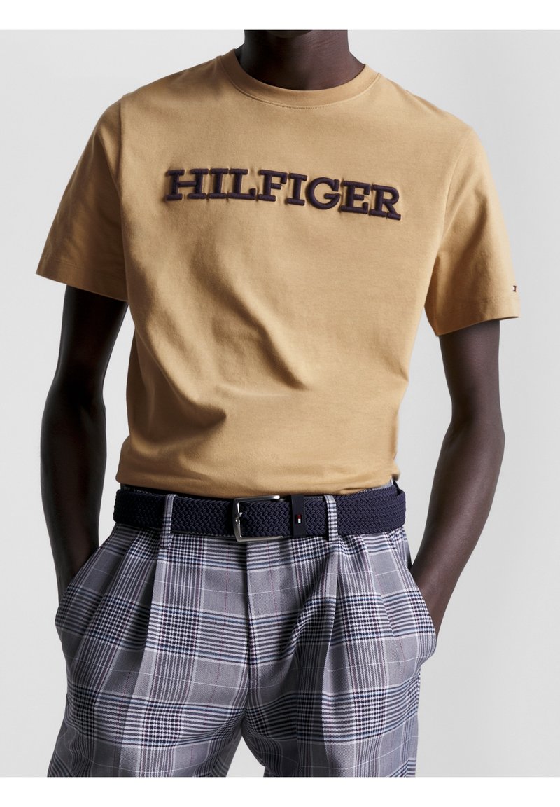 Man som har på sig en beige T-shirt med texten "HILFIGER", rutiga grå byxor och ett marinblått vävt bälte med en silverfärgad spänne, med händerna i fickorna.