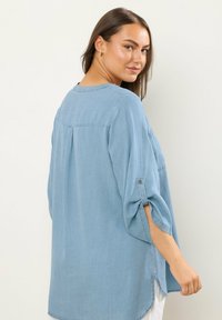 Lys blå denimskjorte med en afslappet pasform, ærmer til albuen, knaplukning ved håndleddene og slidser i siden. Simpelt rygdesign.