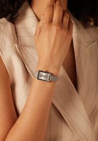 Vierkante zilveren horloge met een grijze siliconenband. De wijzerplaat heeft een zilveren wijzerplaat met verhoogde uurmarkeringen en eenvoudige wijzers.