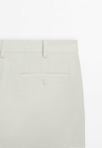 Pantalon beige clair montrant la ceinture arrière avec passants pour ceinture et une poche passepoilée simple avec bouton.