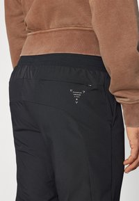 Pantaloni sportivi neri con una finitura testurizzata, dotati di una tasca con zip e dettaglio del logo sul retro, abbinati a un felpa marrone.
