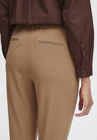 Braunes Langarmshirt kombiniert mit beigen Hosen, die einen elastischen Bund und zwei Gesäßtaschen aufweisen und eine glatte Textur haben.