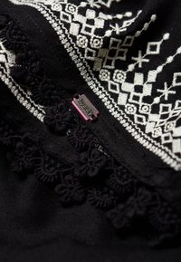 Tissu noir avec broderie géométrique blanche et bordure en dentelle. Une petite étiquette "Superdry" rose est visible près du détail.
