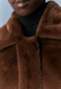 Hnědý kabát z umělé kožešiny s měkkou, hladkou texturou a vysokým límcem, vrstvený přes černý rolák.