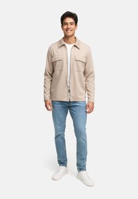 Beige hættejakke med lynlås og to brystlommer, båret over en hvid skjorte. Sammen med lyseblå jeans og hvide sneakers. Glat tekstur.