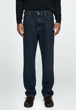 Mand iført mørkeblå jeans med lige ben med knap- og lynlåslukning, kombineret med mørkebrune snørebåndssko og en sort sweater.
