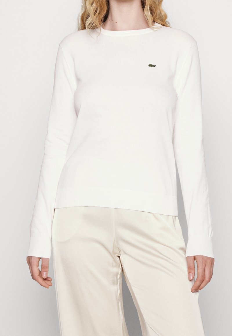 Femme portant un pull blanc à manches longues avec un petit logo crocodile vert et un pantalon beige clair sur un fond uni.