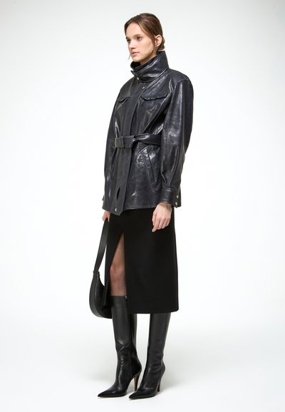 Vrouw draagt een zwarte leren jas met riem, een zwarte midirok met een vooraan gespleten zoom, zwarte leren laarzen tot aan de knie en houdt een zwarte handtas vast.