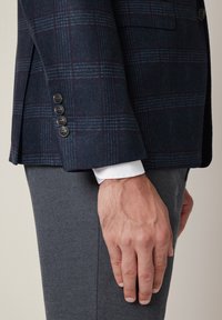 Granatowa marynarka w kratę, wykonana z mieszanki wełny, z burgundowymi akcentami, prezentująca cztery czarne guziki na rękawie i białym mankiecie koszuli pod spodem.