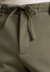 Light olive-green pants made of textured fabric. Features a drawstring waist, front pockets, and subtle stitching details.