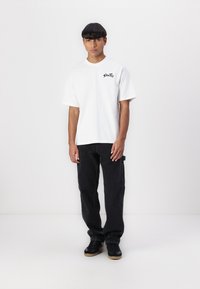 White cotton t-shirt with short sleeves, featuring a black graphic logo. Paired with loose-fitting black jeans and black sneakers.