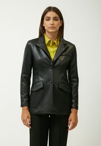 Blazer in pelle nera con design aderente, chiusura a bottone singolo, due tasche frontali e una texture liscia. Indossato sopra una camicia gialla brillante.